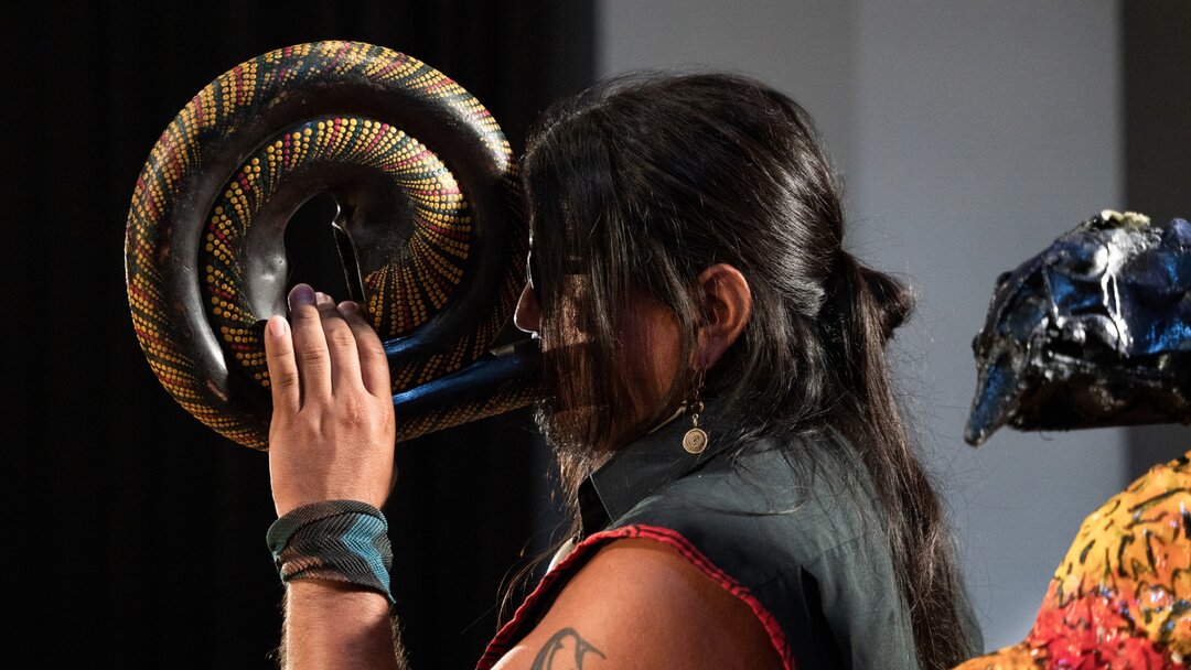 A man in sunglasses and a sleeveless shirt plays a curved horn instrument. Photo by Larissa Matheus.